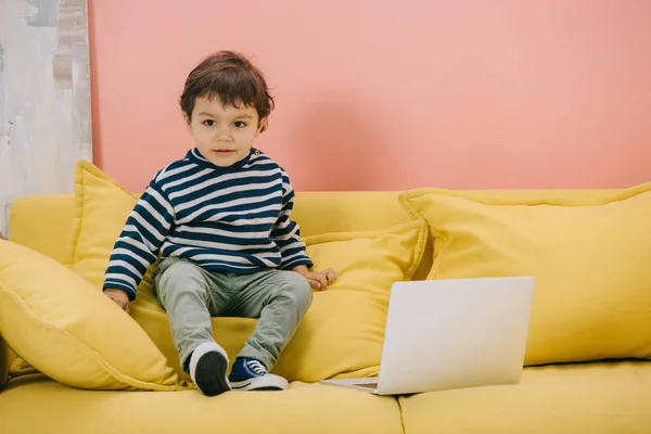 Menino bonito sentado no sofá amarelo perto do laptop — Fotografia de Stock