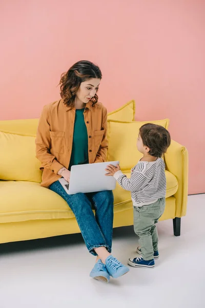 Mother sitting on yellow sofa and using laptop while little son standing near — Stock Photo