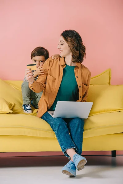 Mãe sorrindo mostrando cartão de crédito para o filho pequeno ao usar laptop — Fotografia de Stock