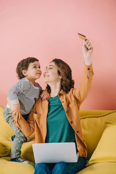 Smiling mother with laptop on knees holding credit card in raised hand and showing it to little son — Stock Photo