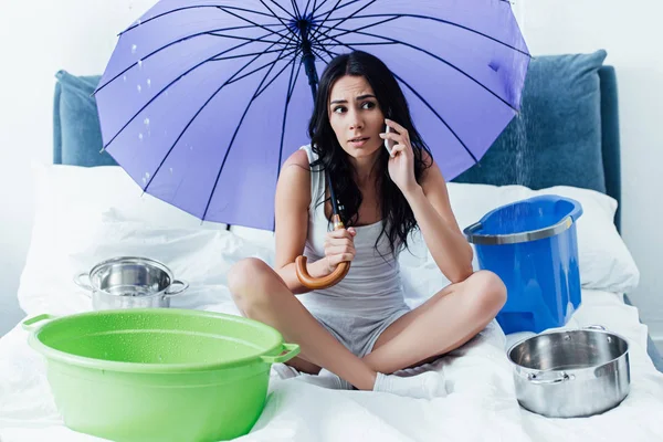 Sad young woman talking on smartphone and holding umbrella in bedroom — Stock Photo