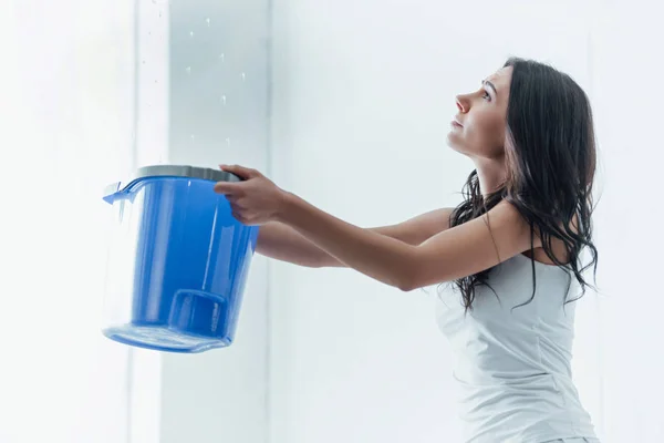 Confus jeune femme tenant seau bleu sous les gouttes d'eau — Photo de stock