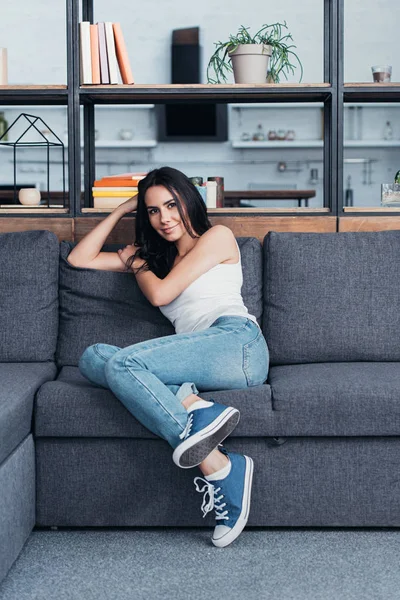 Lächelndes brünettes Mädchen in Jeans sitzt auf Sofa — Stockfoto