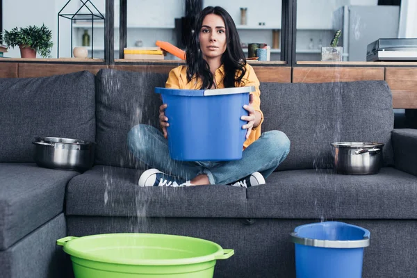 Brünette Frau hält Eimer auf Sofa bei Wasserschaden im Wohnzimmer — Stockfoto