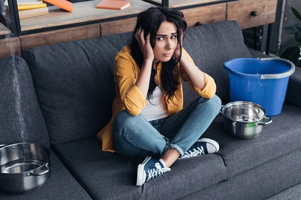 Frustrated brunette woman sitting on sofa under leaking ceiling — Stock Photo