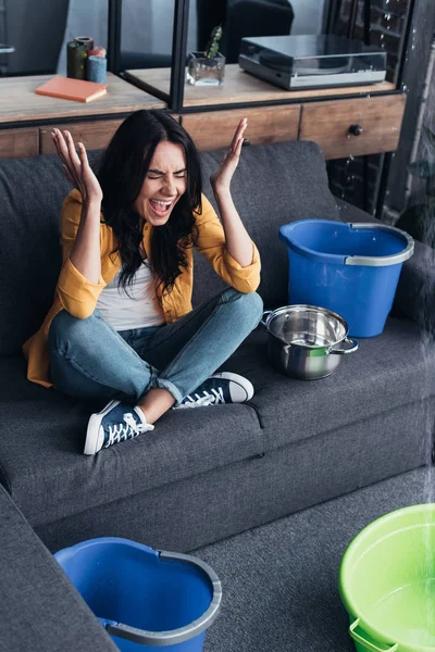 Screaming woman in yellow shirt sitting on sofa under leaking ceiling — Stock Photo