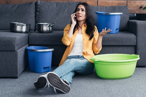 Mujer preocupada hablando en el teléfono inteligente y sentada en el suelo bajo el techo con fugas en la sala de estar - foto de stock