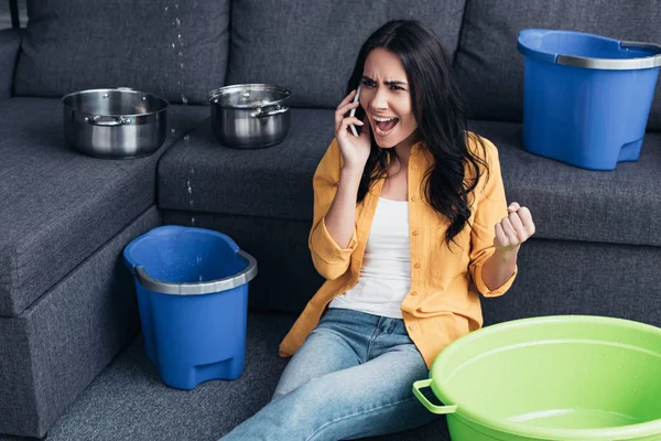 Aggressive woman talking on smartphone and sitting on floor under leaking ceiling — Stock Photo