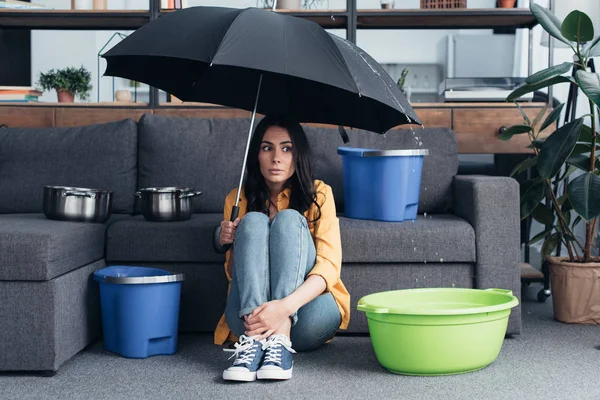 Chica confusa sentada bajo el paraguas en la sala de estar - foto de stock