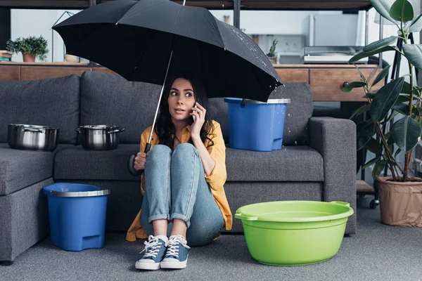 Girl in jeans sitting in living room with umbrella and talking on smartphone — Stock Photo