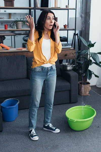 Vista completa de la mujer conmocionada hablando por teléfono mientras el agua gotea del techo - foto de stock
