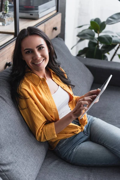 Радісна жінка в жовтій сорочці сидить на дивані з цифровим планшетом — стокове фото
