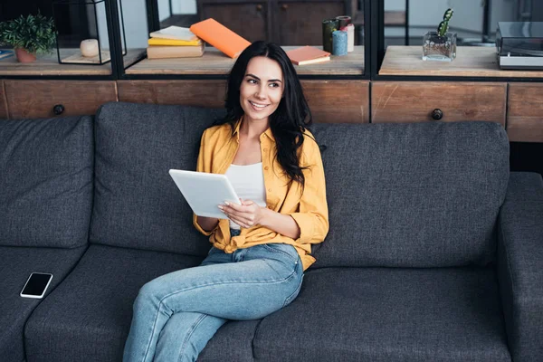 Menina sorridente em camisa amarela sentada no sofá com tablet digital e smartphone com tela em branco — Fotografia de Stock