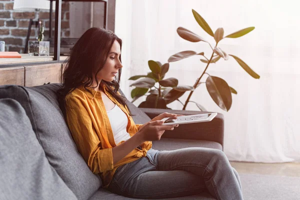 Attractive woman in yellow shirt sitting on sofa with digital tablet — Stock Photo