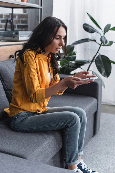 Шокована кучерява дівчина сидить на дивані зі смартфоном — стокове фото