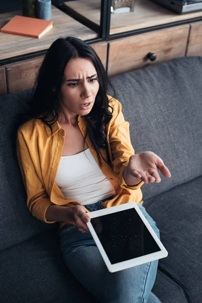 Sconvolto ragazza bruna seduta sul divano e con acqua danneggiato tablet digitale con schermo bianco — Foto stock