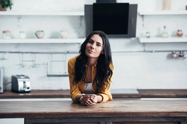 Smiling brunette woman in yellow shirt standing in kitchen with smartphone — Stock Photo