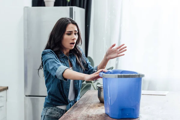 Upset young woman using bucket during leak in kitchen — Stock Photo