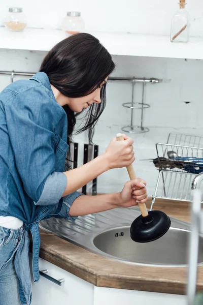 Mujer morena concentrada usando émbolo mientras fija el fregadero - foto de stock