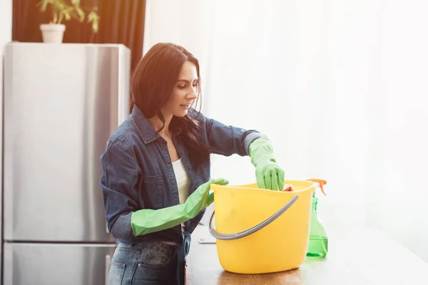 Smiling woman in green rubber gloves using bucket in kitchen — Stock Photo