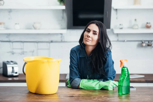 Belle femme brune en gants de caoutchouc assis avec seau et pulvérisation dans la cuisine — Photo de stock