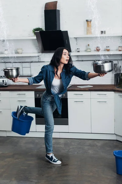 Испуганная молодая женщина с горшками и ведрами, занимающаяся утечкой воды на кухне — стоковое фото
