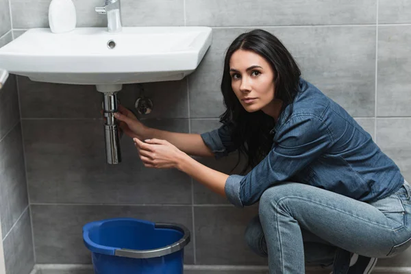 Очаровательная брюнетка в джинсовой рубашке чинит трубу в ванной — стоковое фото