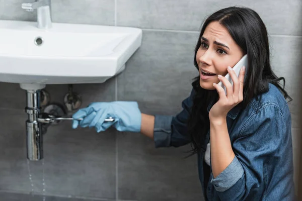 Menina estressada reparação tubo com chave inglesa e falando no smartphone — Fotografia de Stock