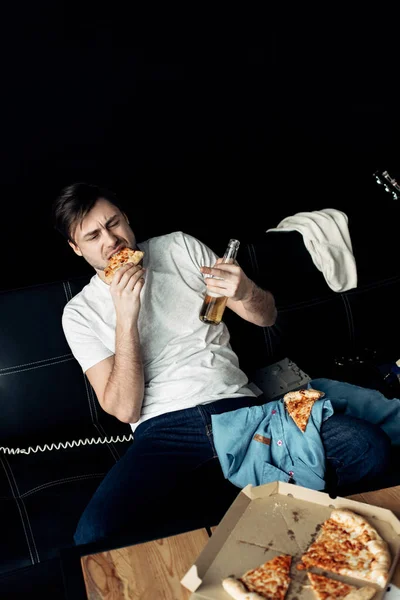 Пьяный человек ест вкусную пиццу и держит бутылку в грязной гостиной — стоковое фото
