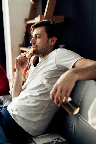 Homem comendo pizza saborosa e segurando garrafa na sala de estar — Fotografia de Stock
