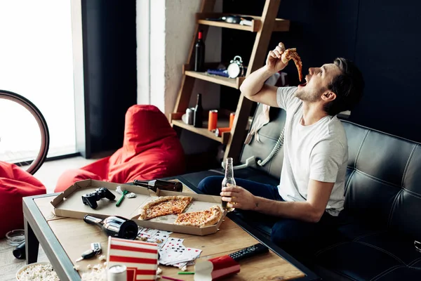 Man eating pizza while holding bottle in messy living room — Stock Photo