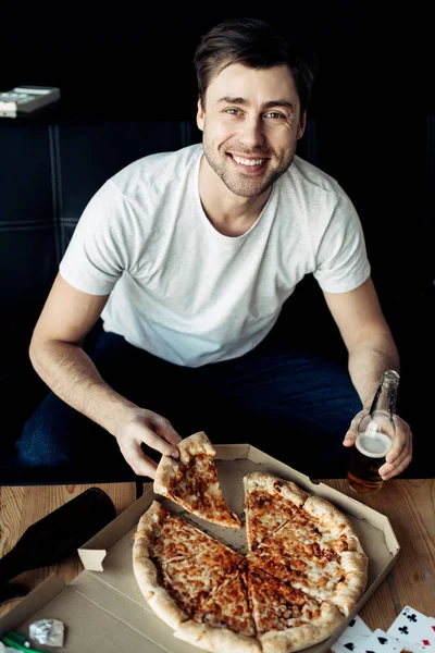 Homem alegre segurando fatia de pizza deliciosa e garrafa na sala de estar — Fotografia de Stock