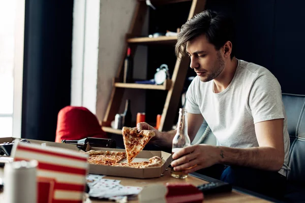 Homem bêbado segurando fatia de pizza deliciosa e garrafa na sala de estar — Fotografia de Stock