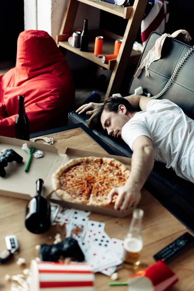 Drunk man sleeping on sofa near tasty pizza at messy home after party — Stock Photo