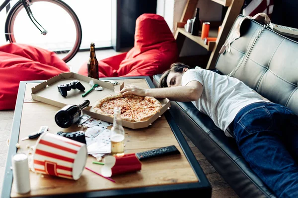 Drunk man lying on sofa near coffee table in messy home after party — Stock Photo