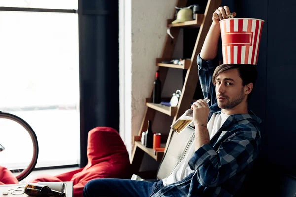 Man holding popcorn box on head drinking beer from bottle at home — Stock Photo