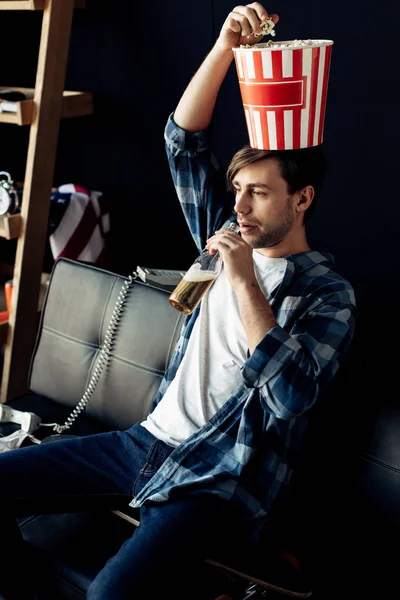 Schöner Mann mit Popcornbox auf dem Kopf trinkt zu Hause Bier aus Flasche — Stockfoto