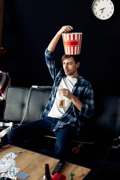 Человек держит коробку попкорна на голове, сидя на диване в грязном доме — стоковое фото