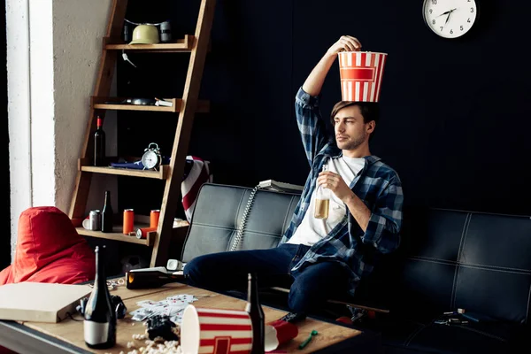 Man holding popcorn box on head and sitting on sofa in messy living room — Stock Photo