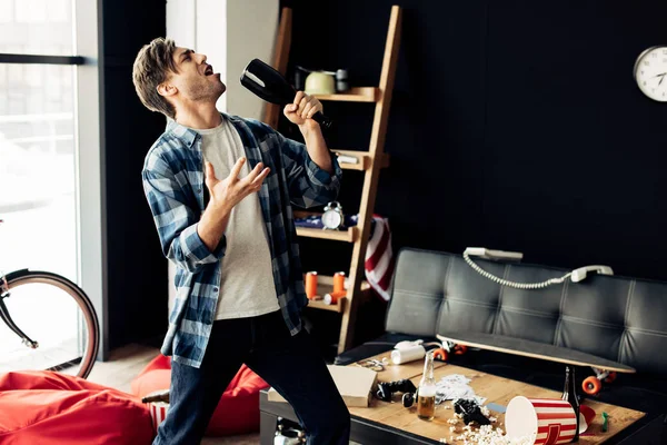 Пьяный мужчина держит бутылку и поет в грязной гостиной после вечеринки — стоковое фото