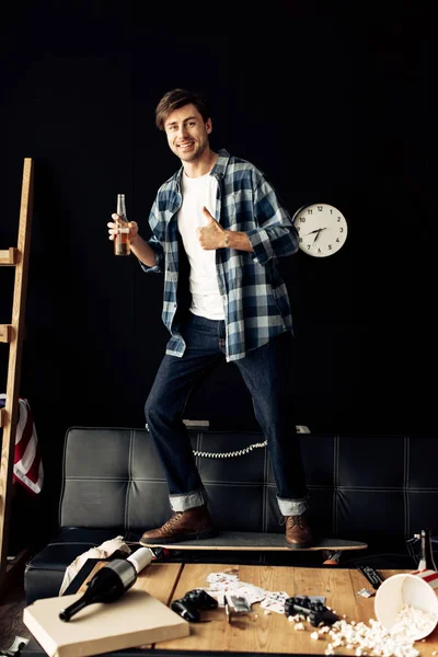 Hombre alegre sosteniendo la botella y mostrando el pulgar en la sala de estar desordenado - foto de stock