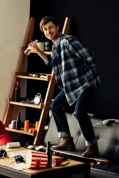 Happy man standing on skateboard on sofa and holding beer in bottle after party — Stock Photo