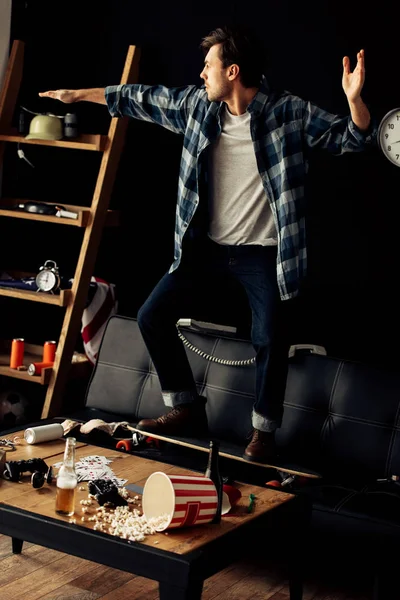 Cheerful man standing on skateboard on sofa and gesturing after party at home — Stock Photo