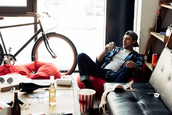 Селективное внимание человека, пьющего пиво из банки и сидящего на бобовом мешке в грязном доме — стоковое фото