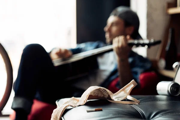 Enfoque selectivo del sujetador cerca del cigarrillo con el guitarrista tocando la guitarra acústica en el fondo - foto de stock