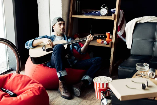 Людина в шапці грає на акустичній гітарі в безладному вітальні — стокове фото