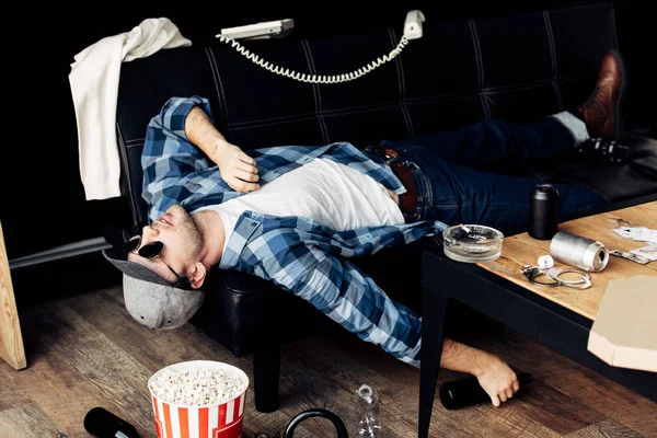 Hombre borracho acostado en el sofá cerca de la botella y la mesa de café en la sala de estar desordenado después de la fiesta - foto de stock