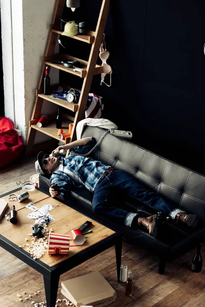 Hombre borracho acostado en el sofá y hablando en el teléfono retro en la sala de estar desordenado después de la fiesta - foto de stock