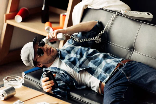 Homem bonito em óculos de sol e boné deitado no sofá e falando no telefone retro na sala de estar — Fotografia de Stock