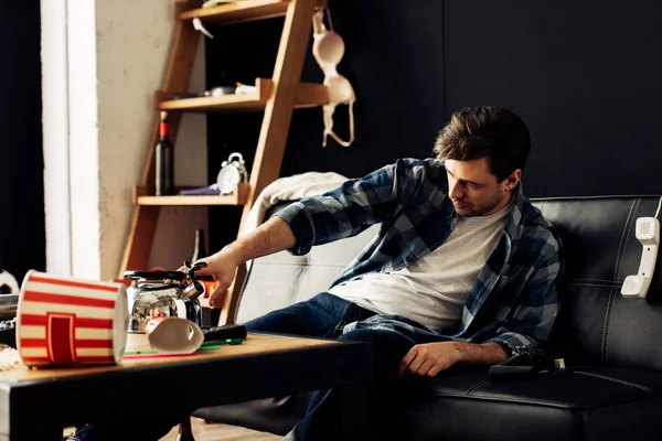 Uomo prendendo teiera da tavolino mentre seduto sul divano in salotto disordinato — Stock Photo